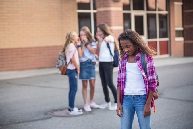 Ein junges Mädchen steht einsam auf dem Schulhof, hinter ihr machen sich drei andere Mädchen über sie lustig.