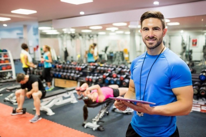 Ein Fitnesstrainer steht zwischen Sportlern in einem Fitnessstudio und hält ein Klemmbrett in der Hand. 