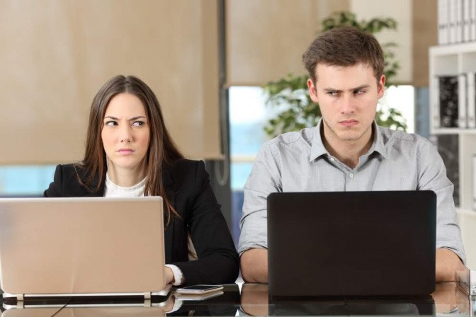 Ein junges Paar sitzt gemeinsam am Schreibtisch, jeder hat einen Laptop vor sich. Sie blicken jedoch beide leicht angenervt schräg hinüber zum jeweils anderen.