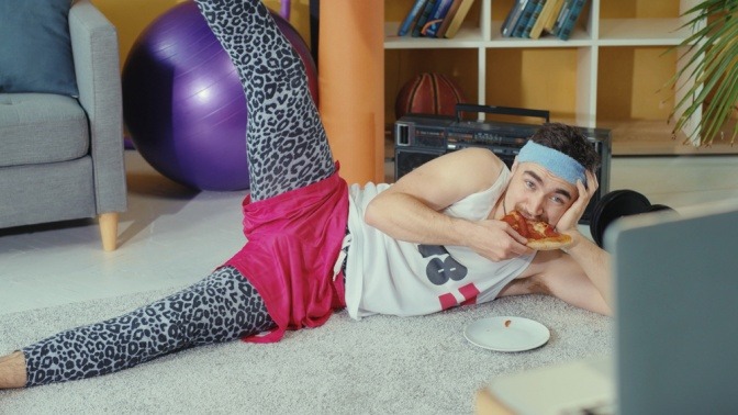 Mann trägt lächerliches Sport-Outfit, bestehend aus Leoparden-Leggins und eine pinke Short darüber, liegt auf dem Boden, ist Pizza und schaut ein Fitness-Video. 