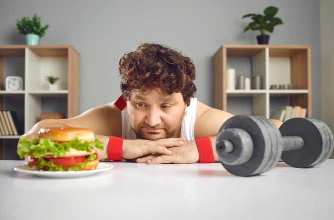 Ein trauriger übergewichtiger Mann im Fitnessoutfit schaut zum Burger statt zur Hantel