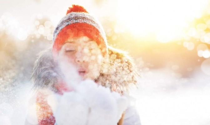 Eine Frau steht draußen im Schnee und pustet eine Wolke mit Pulverschnee von ihren Handflächen.