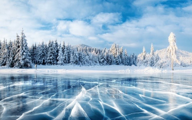 Ein zugefrorener See im Vordergrund, schneebedeckte Nadelbäume im Hintergrund.