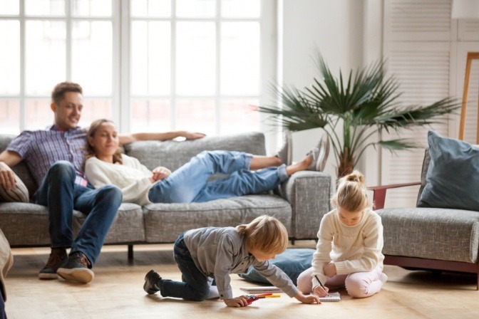 Zwei Kinder spielen auf dem Wohnzimmerboden, während die Eltern auf der Couch entspannen