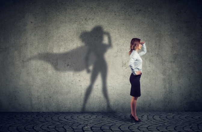 Eine junge Frau steht seitlich zur Kamera und hält nach etwas Ausschau. Ihr Schattenwurf an der Wand gleicht der Silhouette eines Superhelden, samt Umhang und muskelbeladenem Körper.