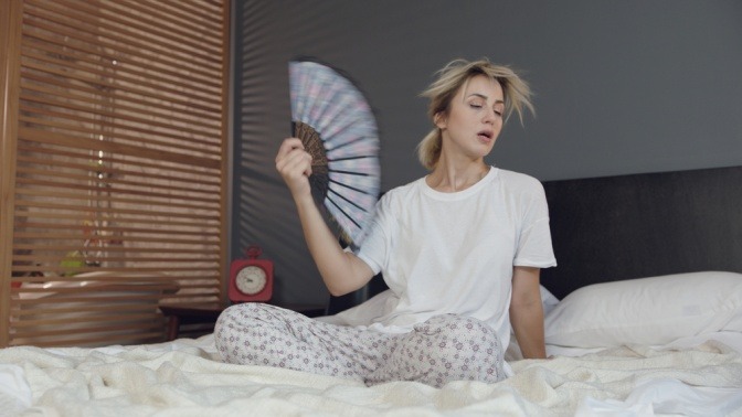 Schwitzende Frau im Bett mit Handfächer. Vielleicht gibt es bei ihr daheim eine zu hohe Luftfeuchtigkeit? Was tun, fragt sie sich jetzt.
