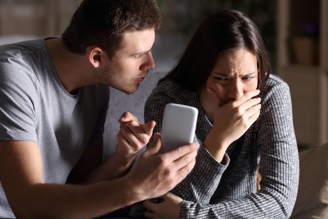 Ein Mann hält seiner Frau vorwurfsvoll ein Handy vor ihr Gesicht, während sie weint. 