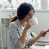 Frau sitzt am Tisch, schaut auf ihr Handy und trinkt dabei gelassen aus einer Tasse.