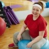 Eine Frau sitzt in einem Sportraum auf einem Gymnastikball und blickt zufrieden in die Kamera.