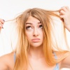 Eine Frau hält Strähnen ihrer blonden Haare in der Hand und schaut verzweifelt. Die Haare sehen sehr trocken aus.