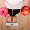 Frau auf Waage mit Doughnut und Apfel in den Händen