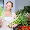 Eine Frau steht in ihrer Küche und hält diverse gesunde Lebensmittel wie Salate und anderes Gemüse in den Händen, mit denen sie gleich gesund kochen wird.