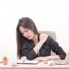 Eine am Schreibtisch sitzende Frau greift sich mit schmerzverzerrtem Gesicht an die Schulter. Rückenschmerzen, Homeoffice und eine fehlende Routine bei der Heimarbeit hängen oft miteinander zusammen.