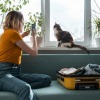 Wohin mit der Katze im Urlaub? Frau macht Foto von Katze auf Sofa neben einem geöffneten Reisekoffer.