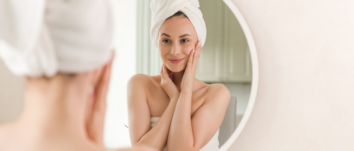 Frau steht nach Feuchtigkeitspflege für die Haut lächelnd vor dem Spiegel