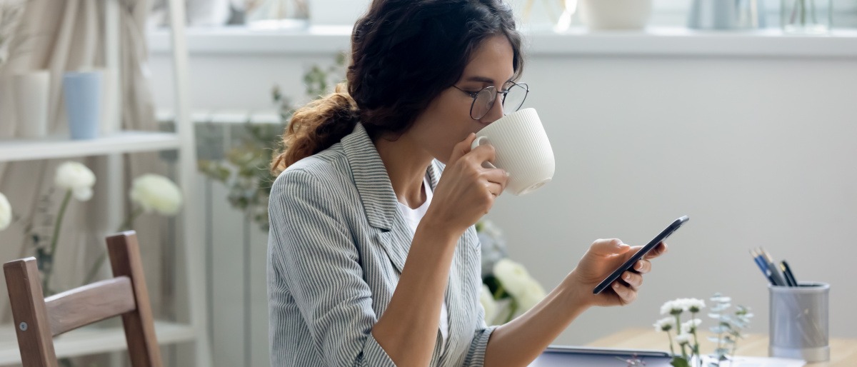 Frau sitzt am Tisch, schaut auf ihr Handy und trinkt dabei gelassen aus einer Tasse.