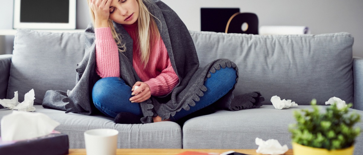 Eine Frau sitzt auf der Couch und greift sich mit der Hand auf die Stirn. Sie fühlt sich sichtlich schwach.