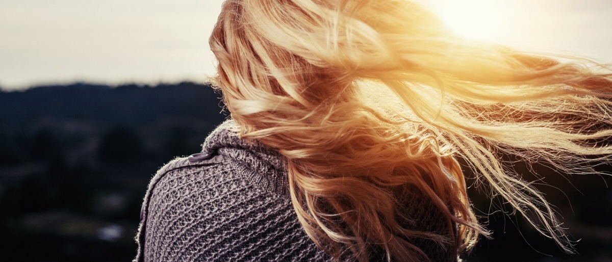 Eine Frau mit prachtvollen, blonden langen Haaren steht mit dem Rücken zum Betrachter gewandt und betrachtet einen Sonnenuntergang, während ihre Haare im Wind fliegen.