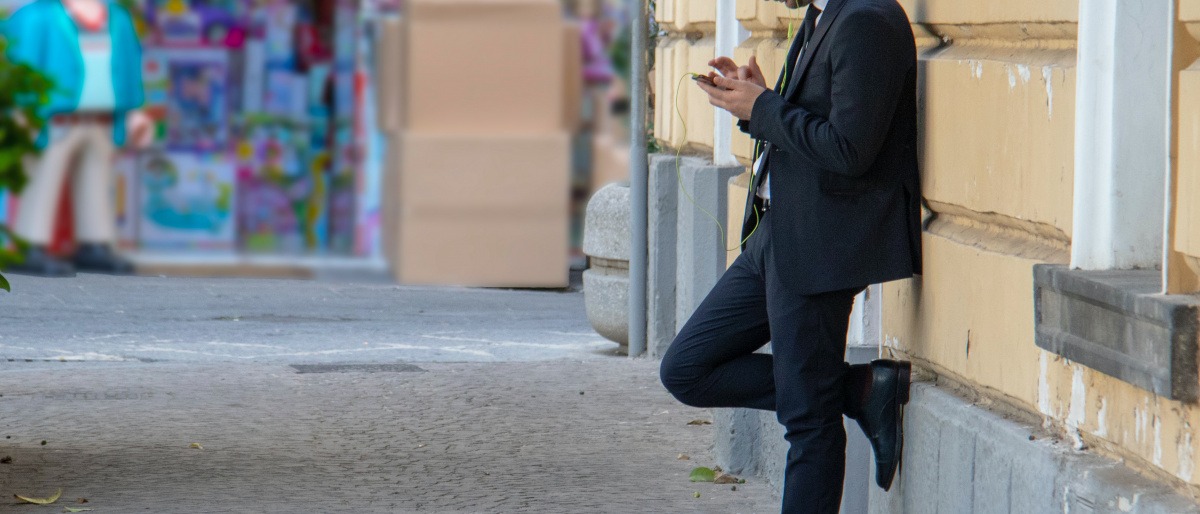 Ein Mann lehnt gedankenverloren an einer Wand und starrt auf sein Handy. 