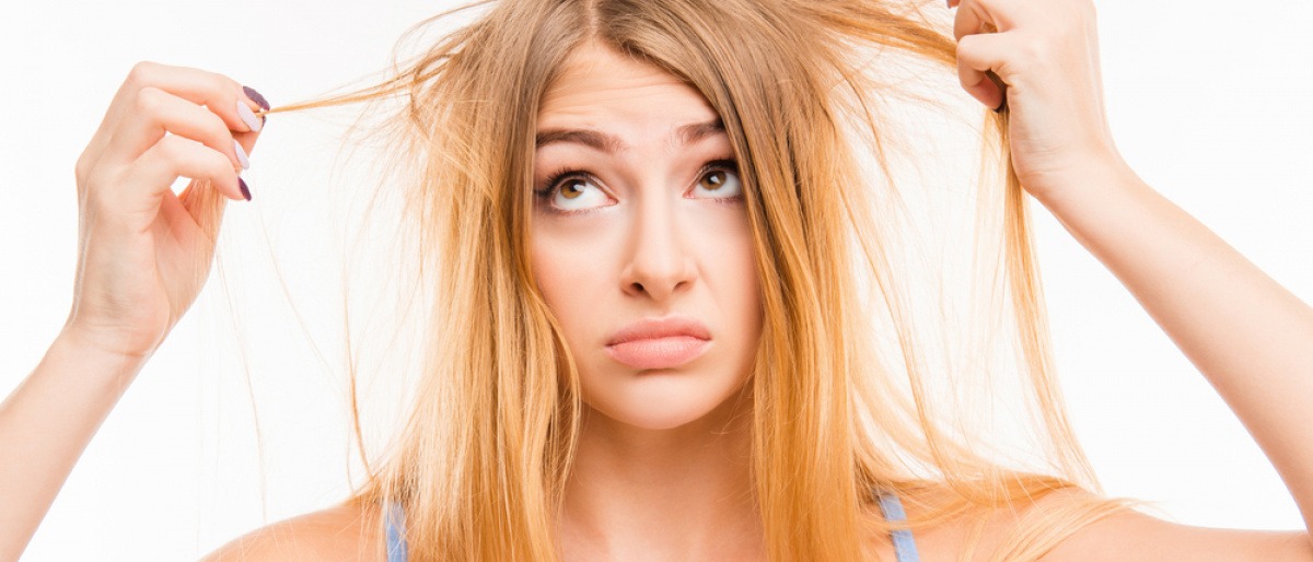 Eine Frau hält Strähnen ihrer blonden Haare in der Hand und schaut verzweifelt. Die Haare sehen sehr trocken aus.