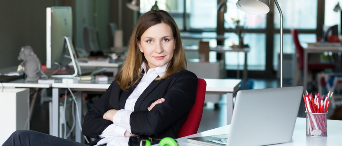 Frau in Businessoutfit sitzt im Cafe und blickt fordernd und selbstbewusst in die Kamera.