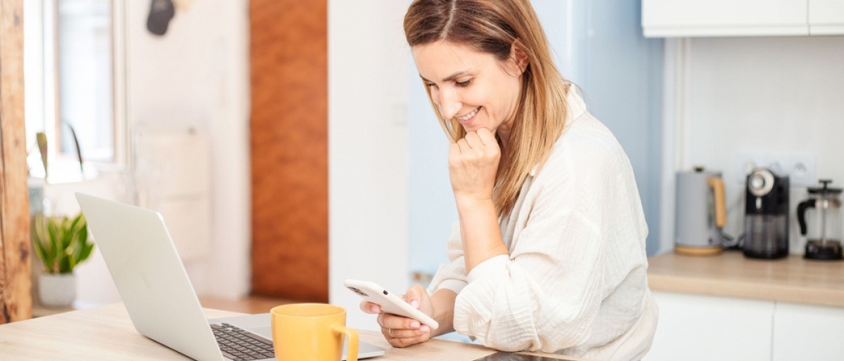 Frau mit Smartphone und Notebook sitzt in Küche mit Elektrogeräten