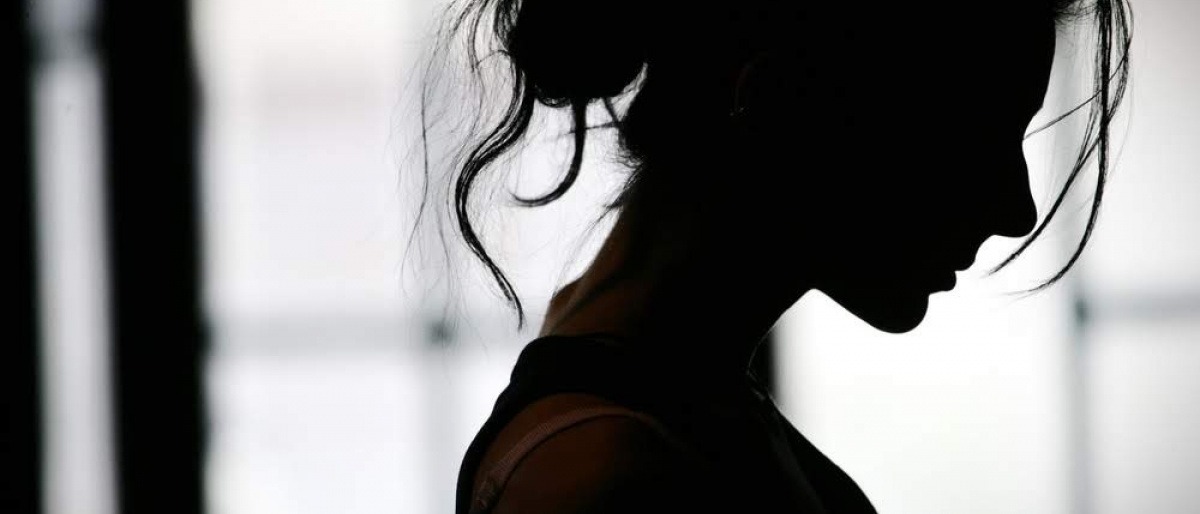 Eine Frau sitzt mit gebeugter Haltung in einer dunklen Atmosphäre hinter einer Milchglaswand. Es wirkt, als müsse sie eine emotionale Krise bewältigen. 