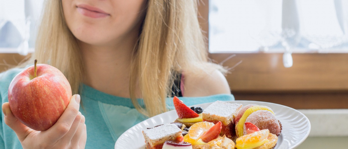 Eine Frau hält einen Apfel und ein Teller mit Süßem und entschließt sich für eine Ernährungsumstellung