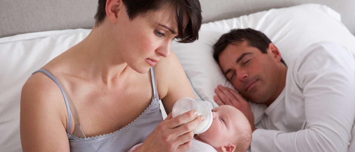 Eine Frau füttert ihr Baby mit der Flasche, der Mann schläft daneben