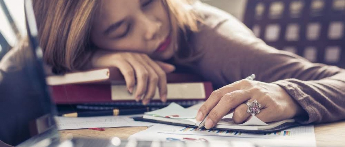 Eine Frau liegt mental erschöpft am Schreibtisch über Büchern und Unterlagen