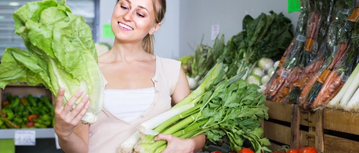 Eine Frau steht in ihrer Küche und hält diverse gesunde Lebensmittel wie Salate und anderes Gemüse in den Händen, mit denen sie gleich gesund kochen wird.