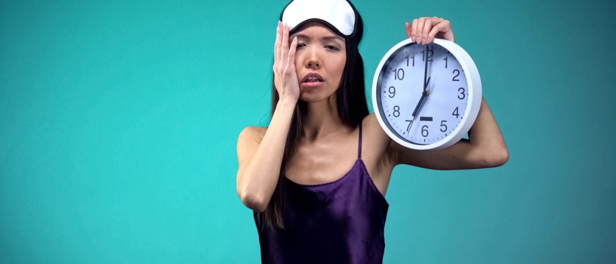 Innere Uhr umstellen - müde wirkende Frau im Nachthemd hält eine Wanduhr hoch.