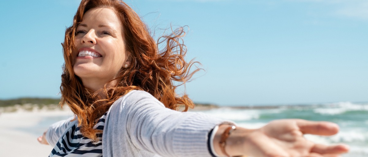 Eine fröhliche reife Frau mit roten Haaren streckt ihre Hände aus und genießt die Strandbrise