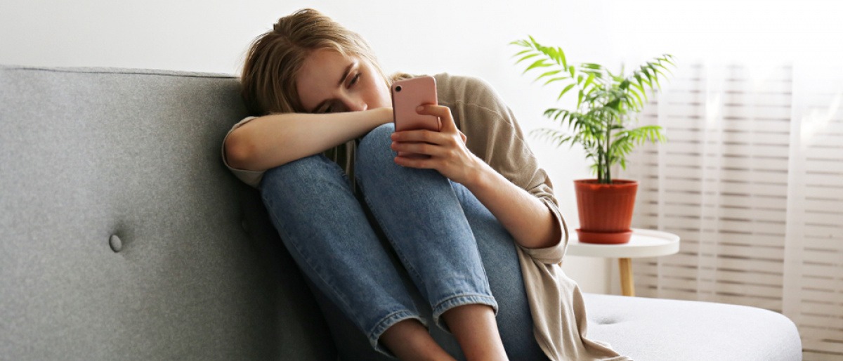 Eine Frau sitzt in zusammengezogener Körperhaltung auf der Couch und schaut traurig in ihr Handy