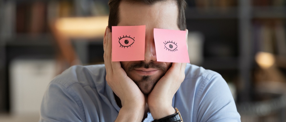 Mann zeigt keine Gefühle - ein Mann sitzt apathisch am Tisch, über seinen Augen kleben rosafarbene Post-its, auf denen Augen aufgemalt sind.
