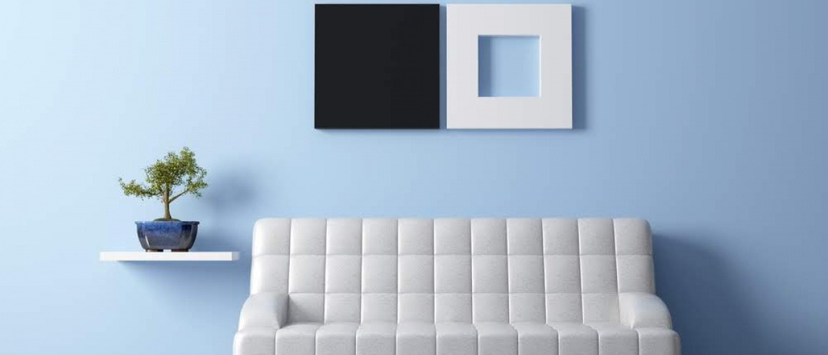 Das Thema minimalistisch Wohnen wird in diesem Bild beispielhaft anhand eines Raums gezeigt, in dem die Einrichtung nur aus einem Sofa, einem Regal mit einer Topfpflanze und einem Bild an der Wand gibt. Der Raum wirkt dennoch einladend und gemütlich.