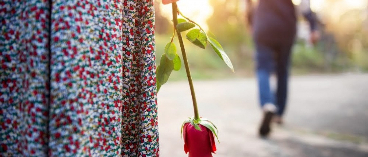 Eine Frau hält eine Rose in der am Körper anliegenden Hand, während ihr Partner sich von ihr weggeht.
