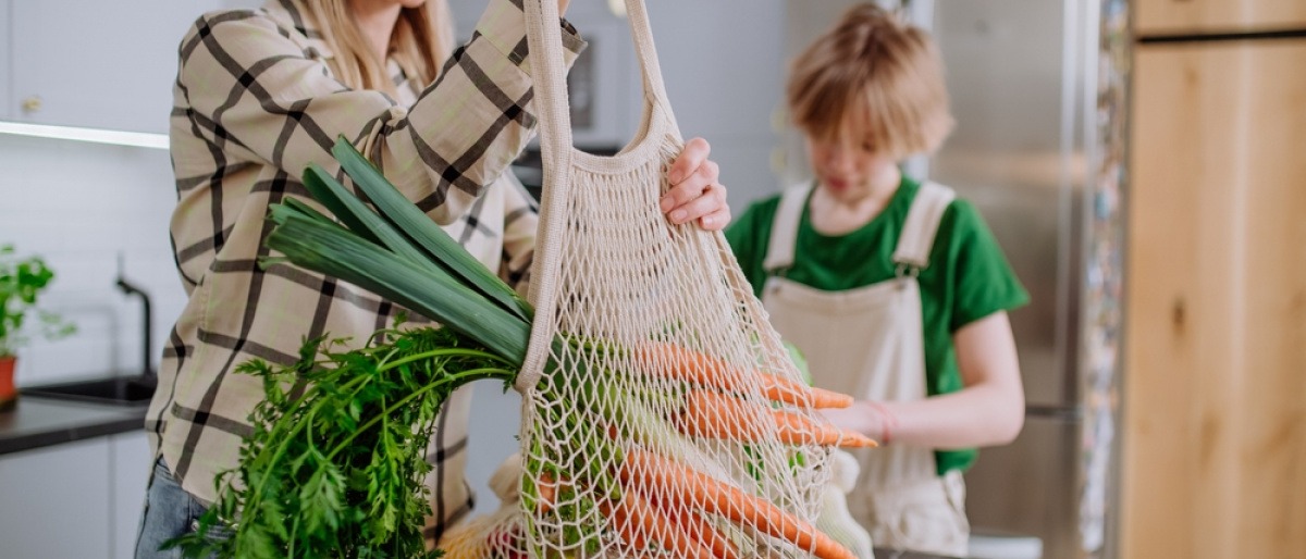 Eine junge Mutter bringt ihr eingekauftes Obst und Gemüse in einem Jutebeutel in die Küche und packt sie gemeinsam mit ihrem Sohn aus.