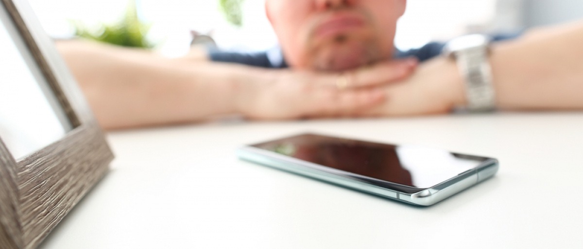 Ein Handy liegt auf dem Tisch. Ein nachdenklicher Mann im Hintergrund wartet auf einen Anruf oder eine Nachricht einer Frau.