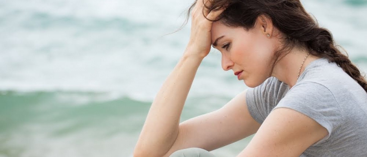 Eine Frau sitzt auf einer Düne am Strand, der Kopf ist nach unten gesenkt, ihr Ausdruck wirkt traurig und ratlos.