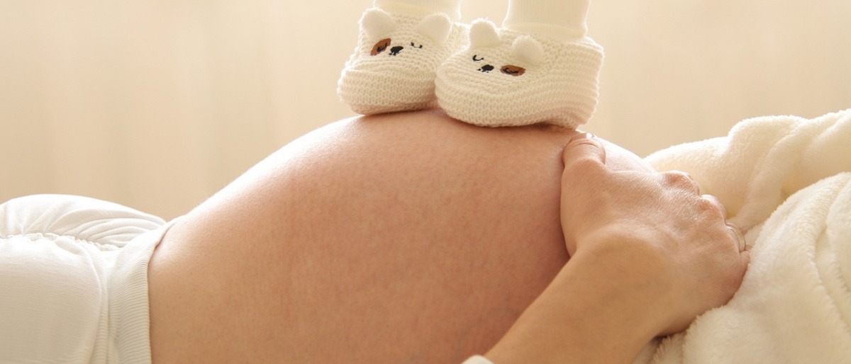 Am Bauch einer schwangeren Frau stehen zwei kleine Schuhe