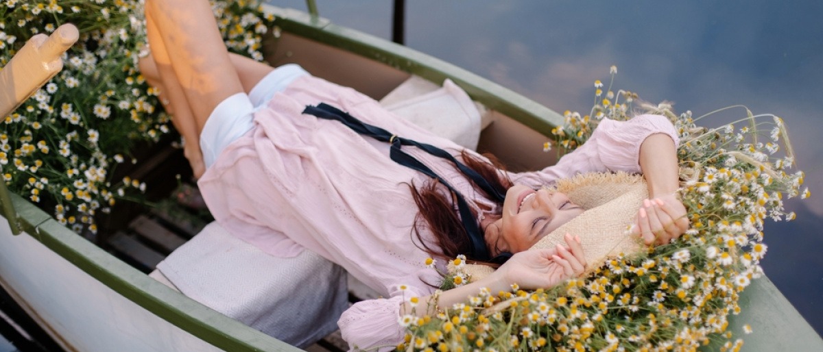 Frau mit Hut liegt im Sommer in einem mit Blumen ausgelegten Boot und lässt sich treiben.