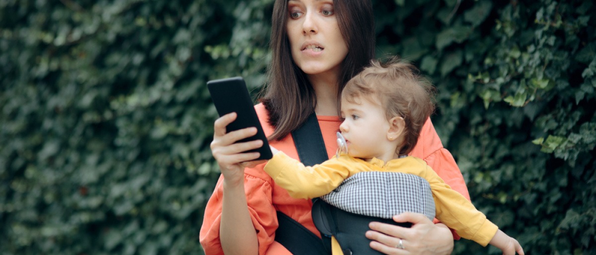 Stress durch Handy - Mutter mit Baby im Arm blickt gestresst auf ihr Smartphone.