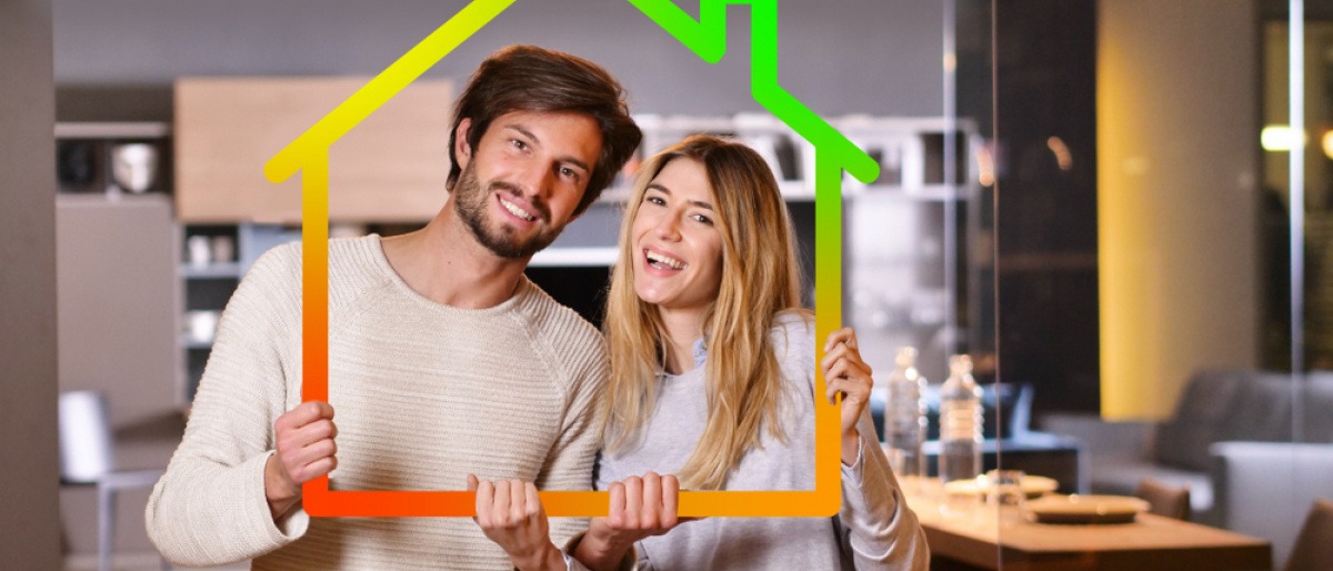 Strom sparen zuhause - junges Paar hält ein kleines Haus-Symbol hoch, dessen Umrisse in grün, gelb und rot gefärbt sind.