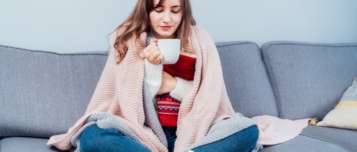 Wärmekissen selber machen - Frau sitzt mit Wärmekissen und Decke über den Schultern auf ihrem Sofa und trinkt Tee.