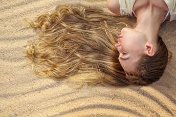 Frau mit langen Haaren liegt im Sand und lässt die Haare von der Sonne aufhellen
