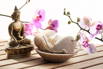 Auf dem Foto ist eine Buddha-Figur abgebildet, davor steht ein Steinschälchen. Im Hintergrund befinden sich Orchideen. 
