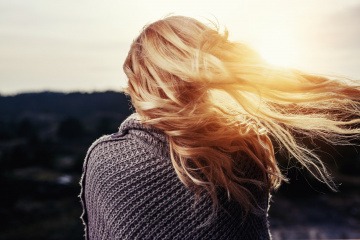 Eine Frau mit prachtvollen, blonden langen Haaren steht mit dem Rücken zum Betrachter gewandt und betrachtet einen Sonnenuntergang, während ihre Haare im Wind fliegen.