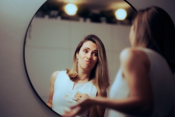 Frau mit Fleck auf weißem Kleid steht vor Spiegel im Badezimmer und wirkt verzweifelt.