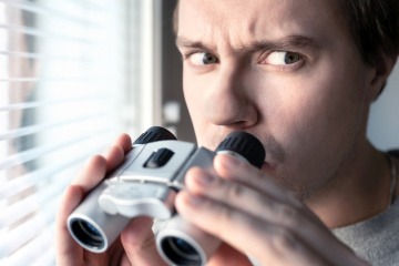 Ein Mann steht mit einem Fernglas hinter einem Fenster und hat einen misstrauischen Blick.